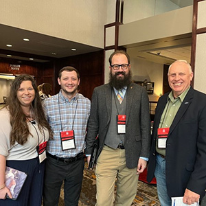 A woman and three men smile for a group photo in a conference room.