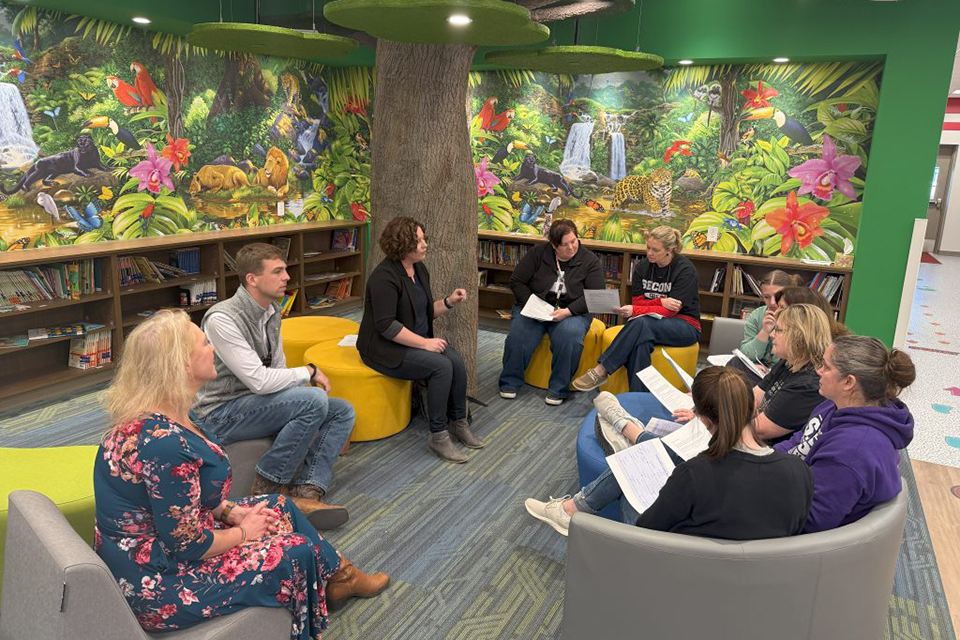 A group of casually dressed researchers meet with the director of a multigenerational community center in a colorful reading corner.