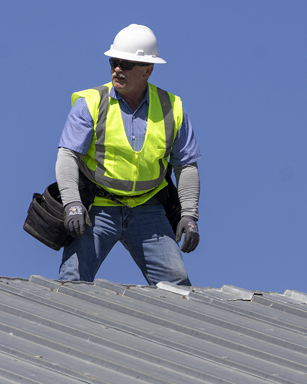 A construction worker in a bright yellow safety vest and white helmet stands atop a metal roof he is helping dismantle.
