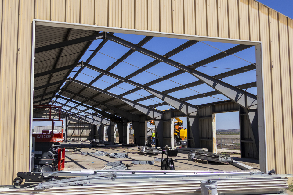 From outside the structure, the camera shoots down a pre-engineered metal structure that is being dismantled, starting with the roof. Bright blue sky peeks through the bare girders on half the roof.