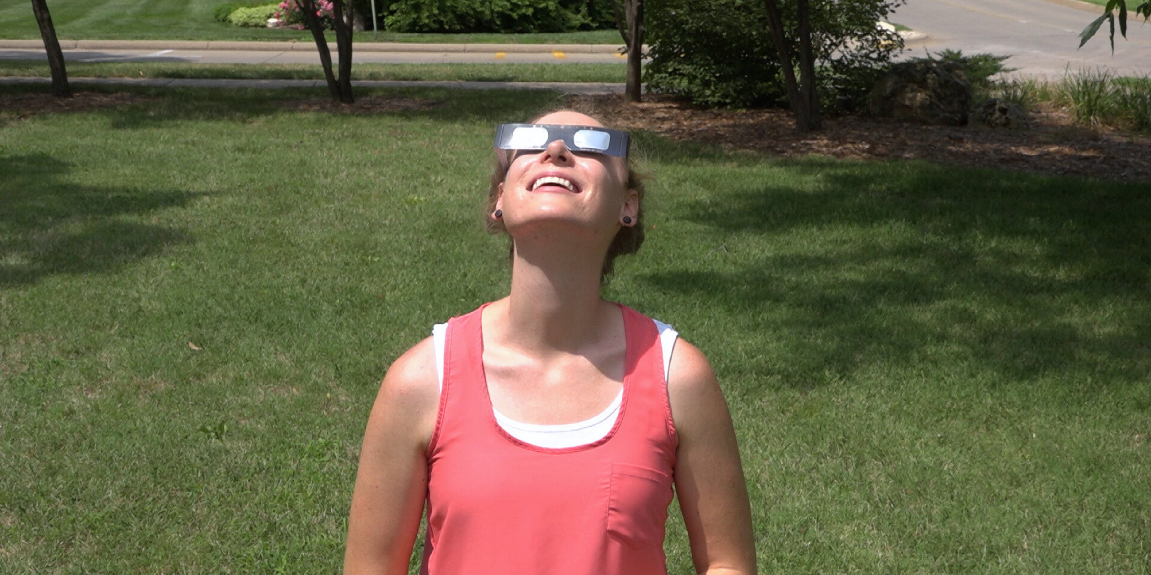 A female student, wearing a coral tank top, looks up at the solar eclipse using proper protective sunglasses.