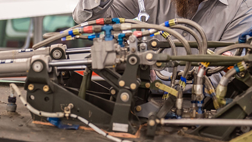 Two men in ballcaps and work shirts hover over the open engine compartment of a propeller airplane in a maintenance hangar. The one on the right holds a rag and an oil dipstick, while the other main points to the part of the dipstick that shows the airplane's oil level.