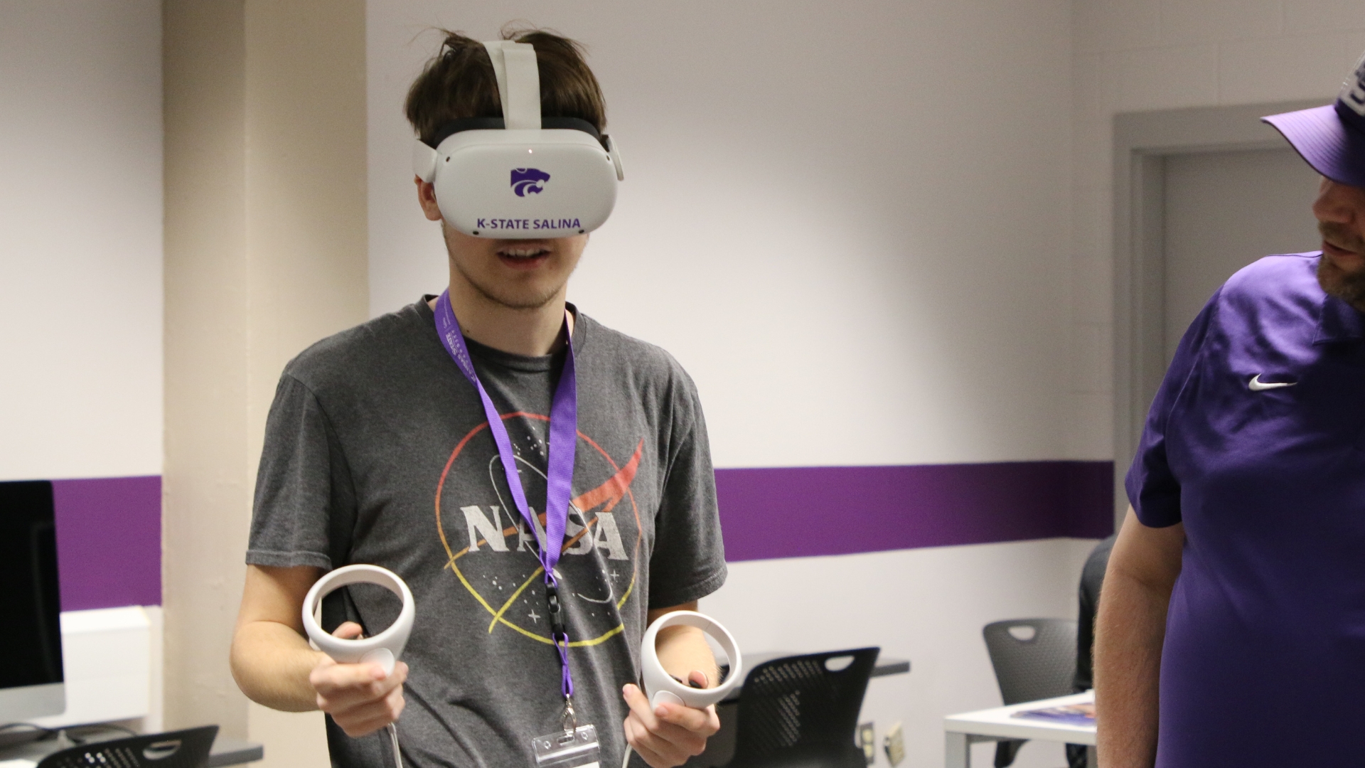 A student in a T-shirt wears white virtual reality goggles as they hold paddles to manipulate a virtual setting.