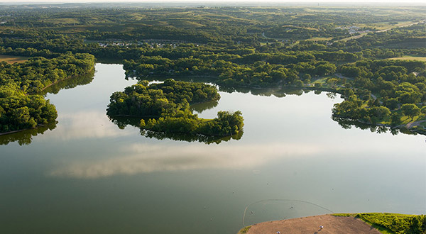 An aerial view shows a snaking dammed lake among a cluster of tree-topped, green hills.