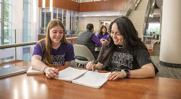 Two students study together at a wooden table in a large, brightly lit atrium.
