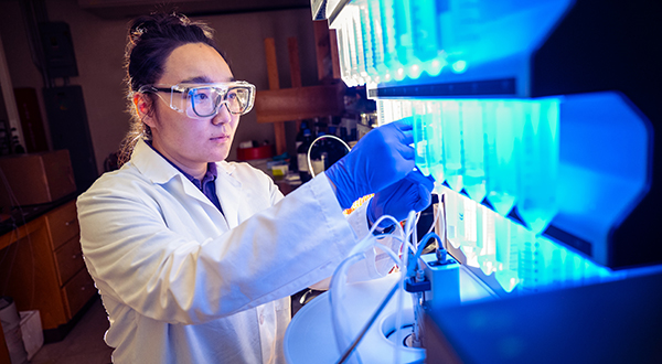 A researcher in a white lab coat works with some bright blue-lit test tubes on a rack.