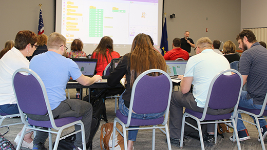 A group of several teacher sits at a table listening to a preentation in a larger conference room.