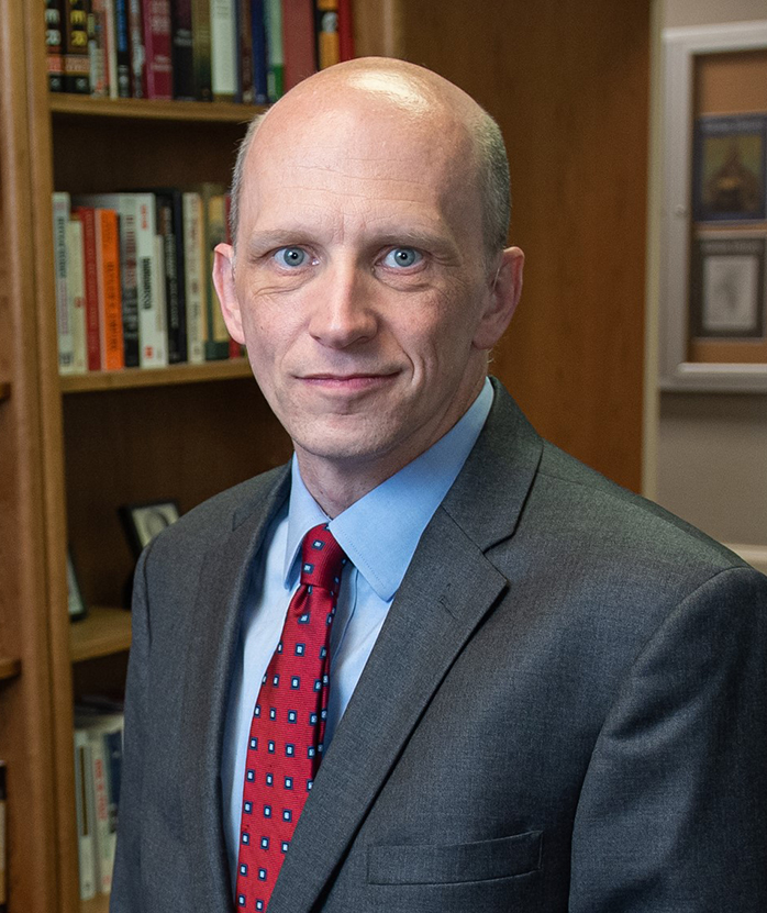 A man in a gray suit, blue shirt and red tie smiles for a portrait photo in front of a bookshelf.