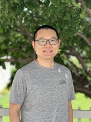A student in glasses and a gray t-shirt stands in front of a fence and a green tree.