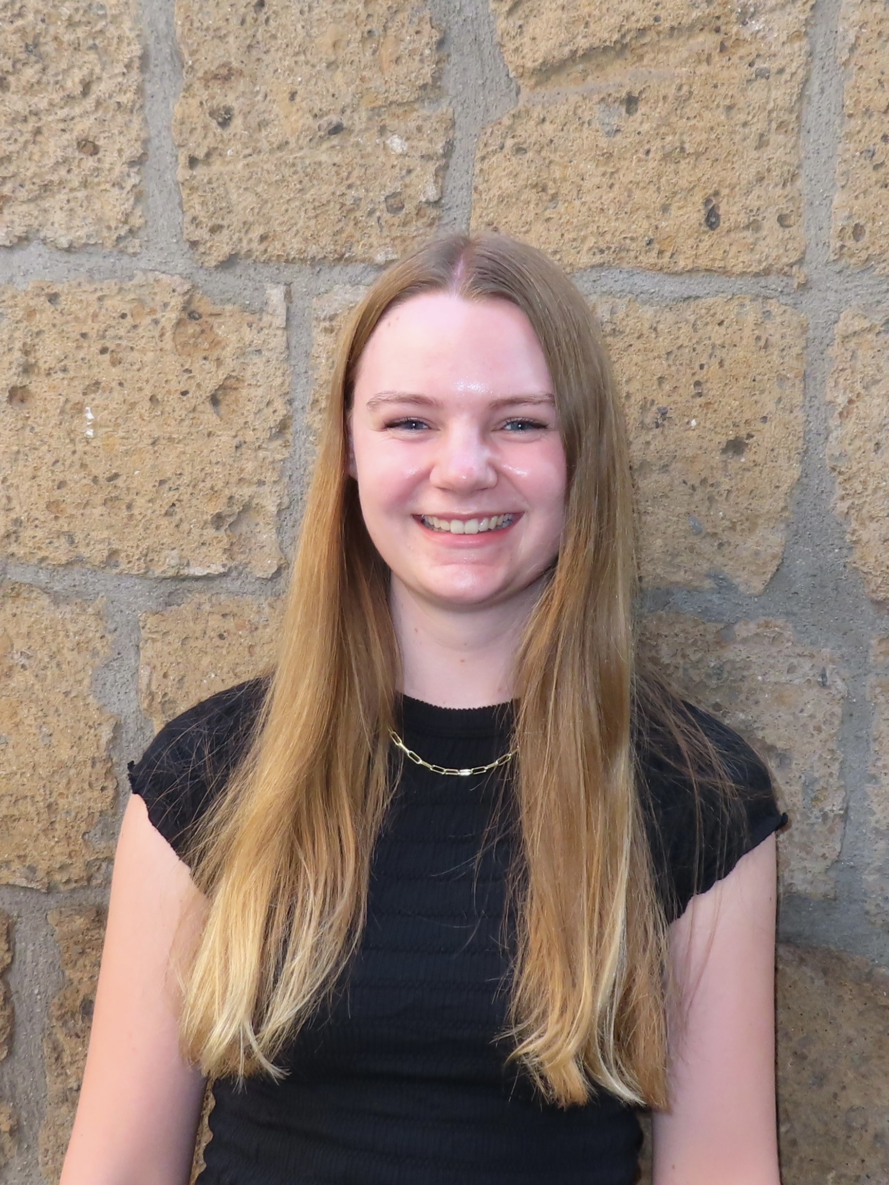 A young woman in a black T-shirt stands in front of a brick wall and smiles for a photo.