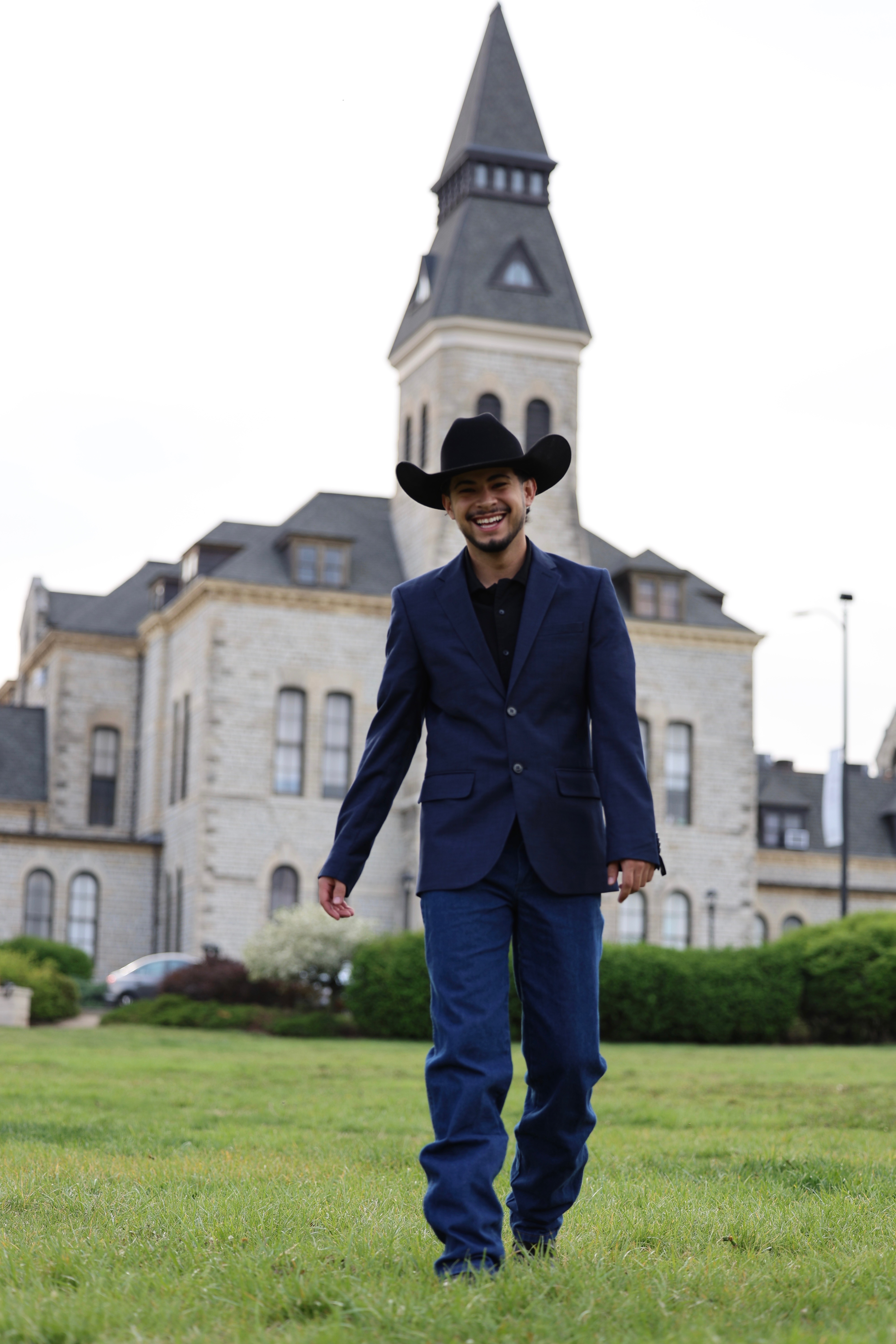A man in jeans and a navy suit jacket wearing a black cowboy hat smiles and walks across the green grass of Anderson Lawn in front of Anderson Hall.