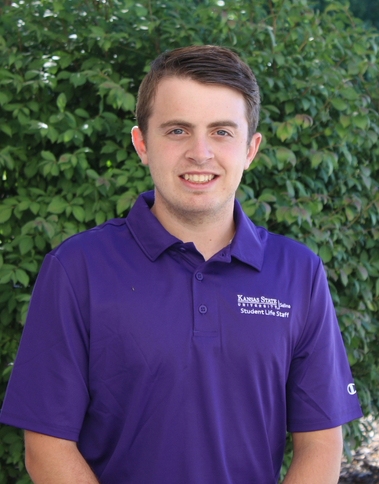 A young man in a purple polo smiles in front of greenery.