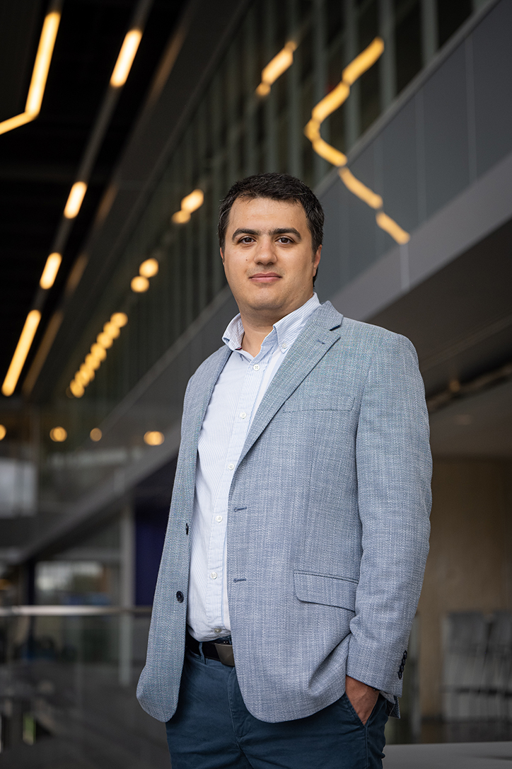 A man in a gray suit and light shirt stands in a building with lights overhead and smiles for a portrait photo.