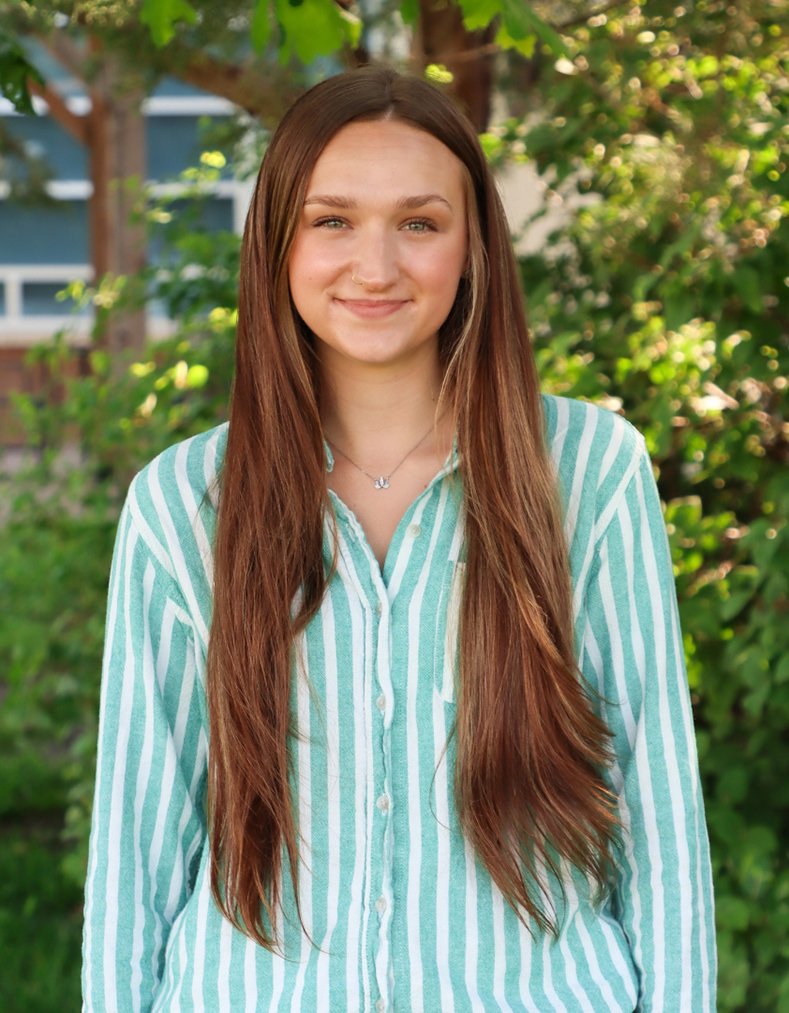 A young woman in a green and white striped shirt smiles in front of greenery.