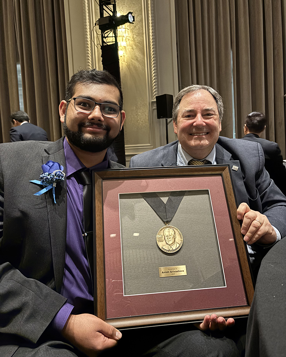 A student and a professor in ties hold up a plaque the student won.