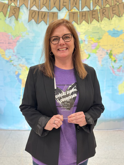 A professor in a blazer holds up a glass award in front of a map background.