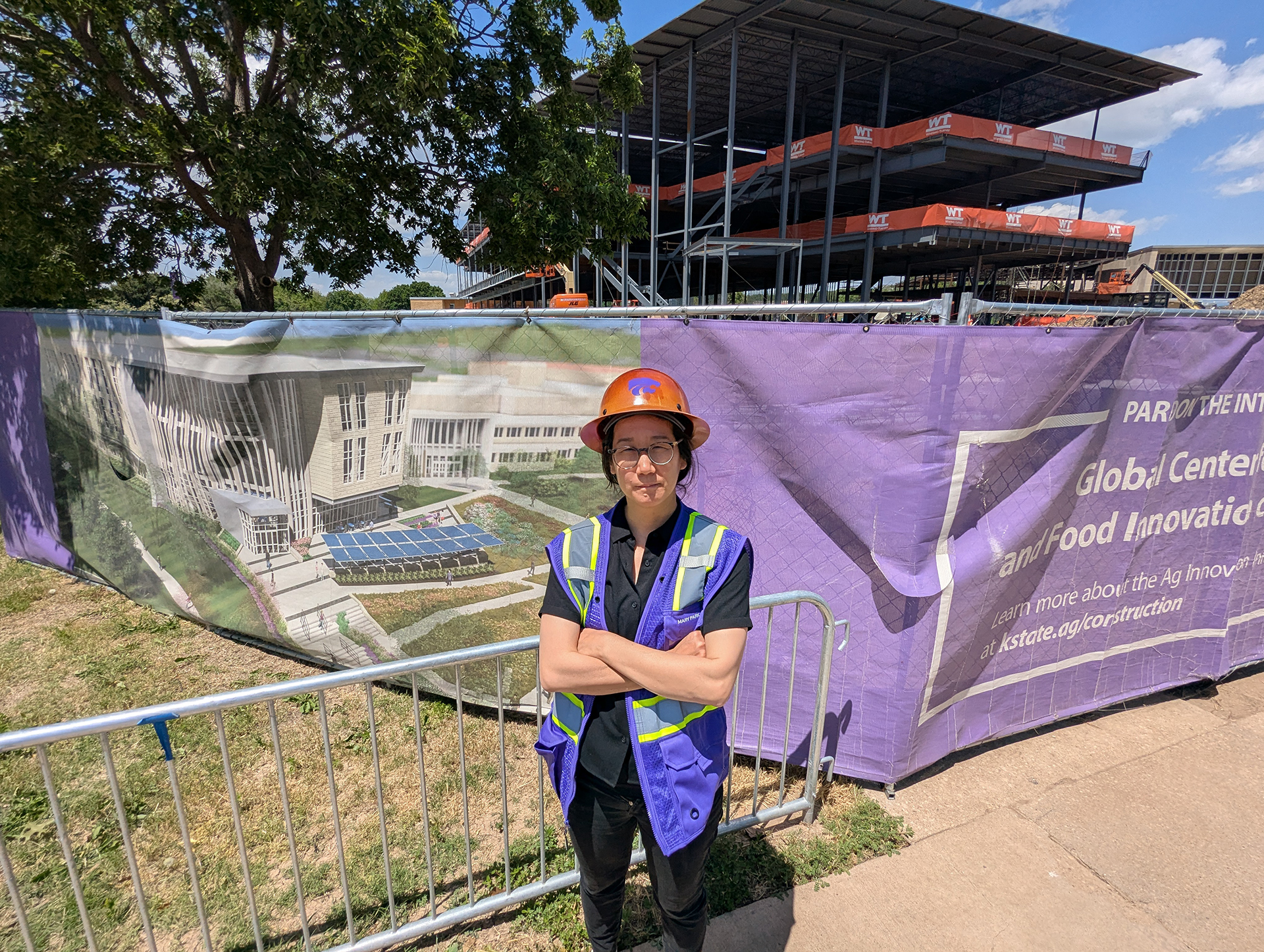 A woman in safety vest stands in front of construction fence