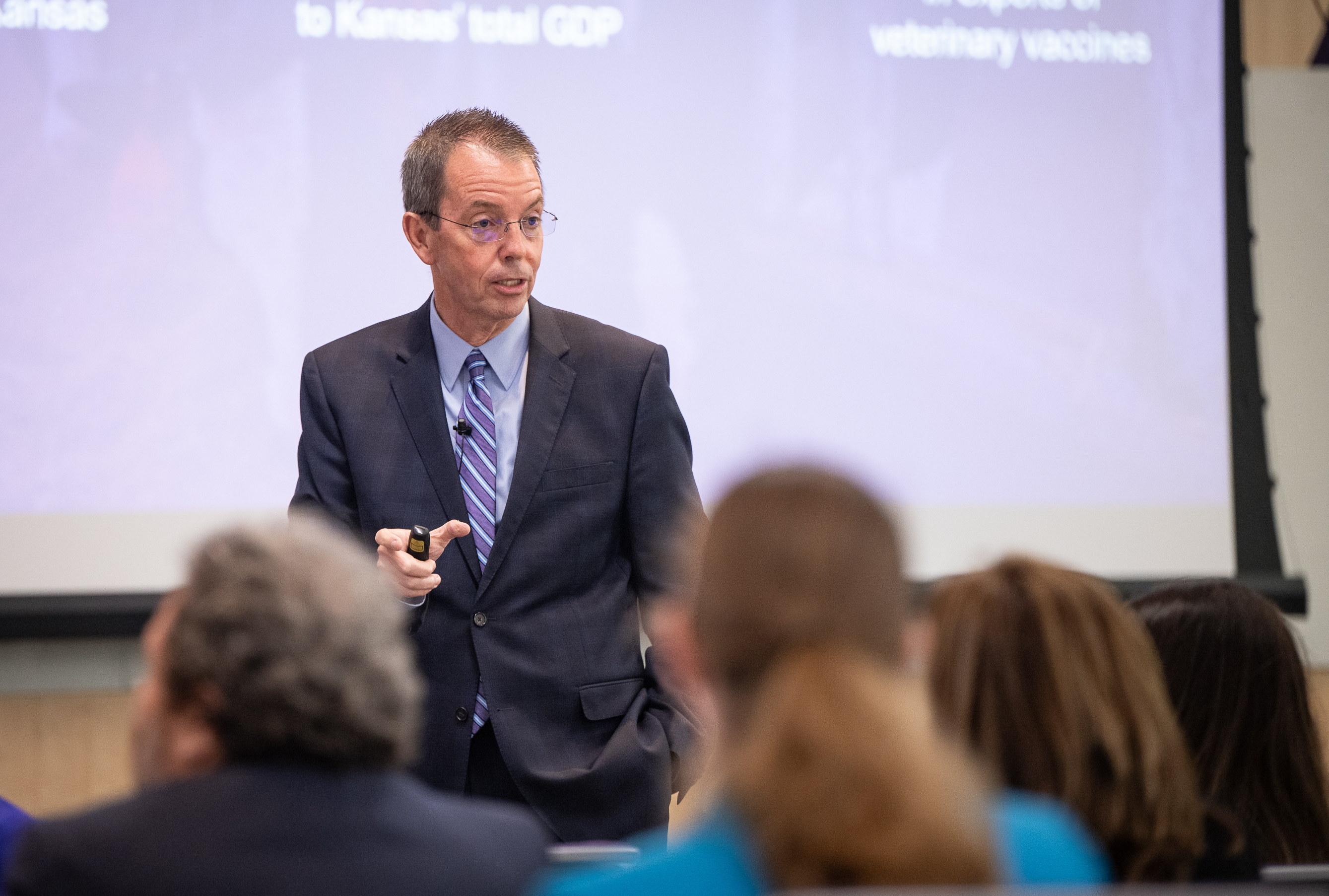 A man in a gray suit and a purple shirt with glasses presents at the front of a room.