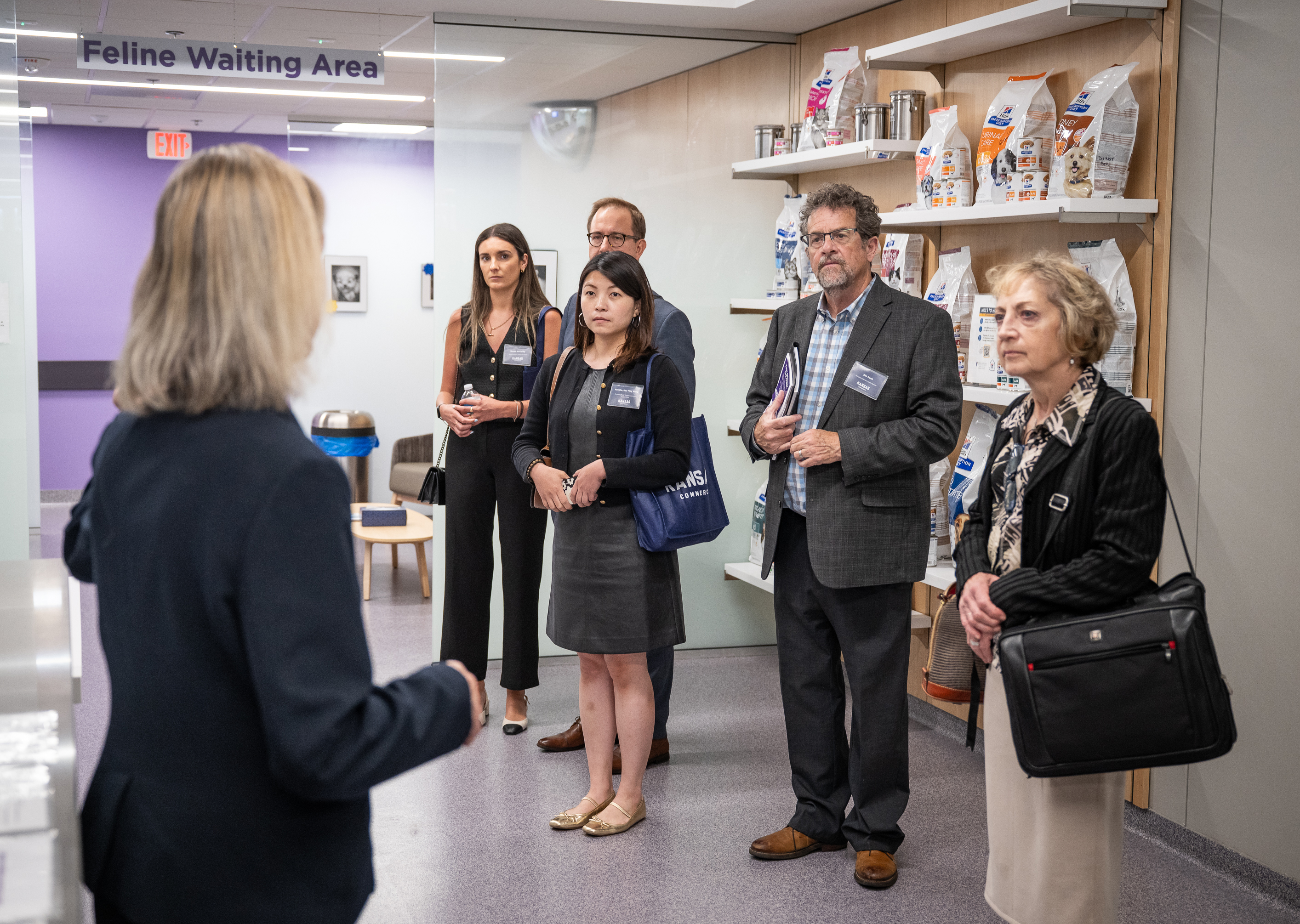 A group of people in business attire stand in front of a shelf of pet food in an area labeled feline waiting room while a person in a suit with their back to the camera presents.