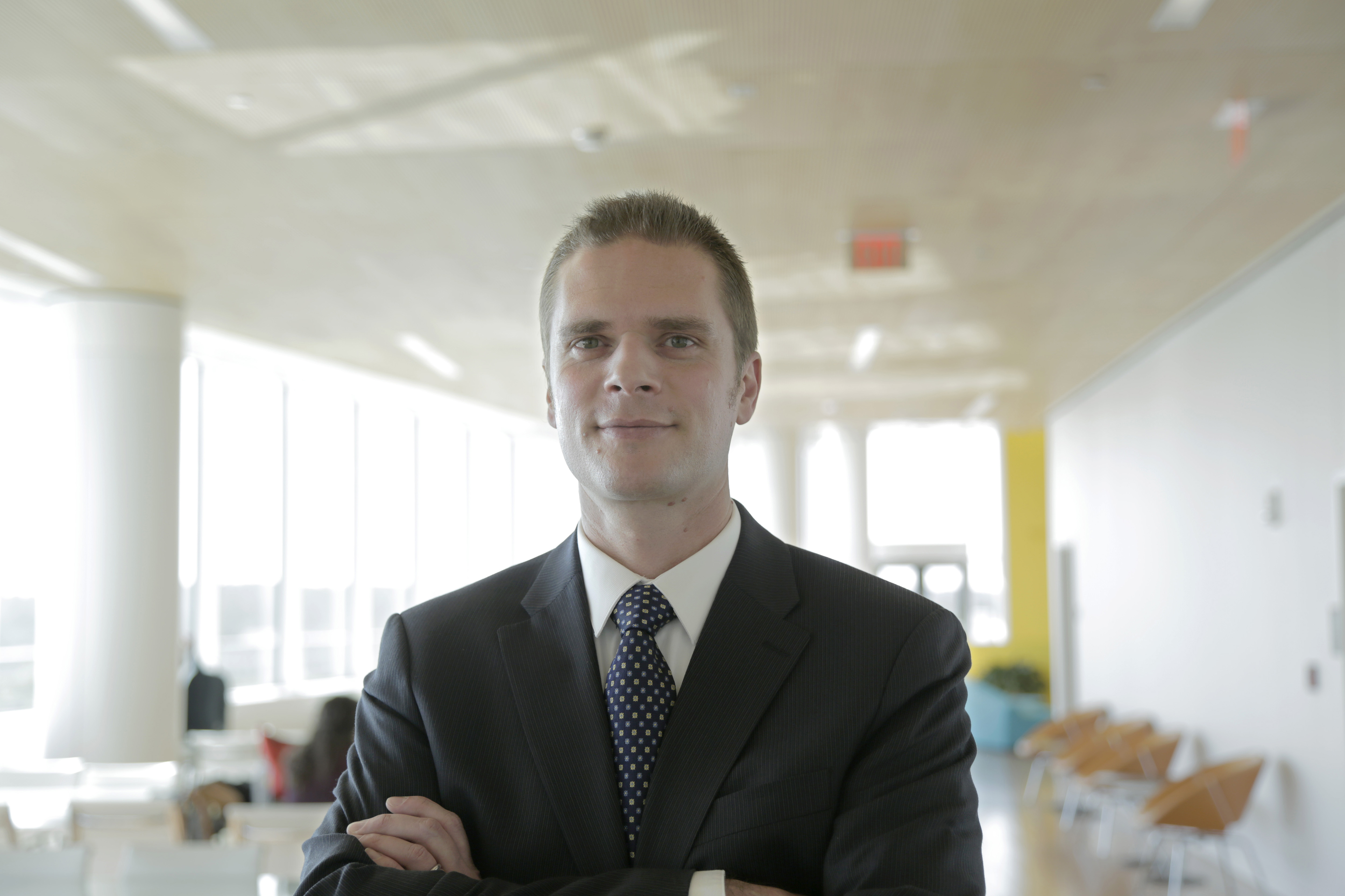 A man in a suit crossing his arms and smiling for a portrait photo in an office with many windows