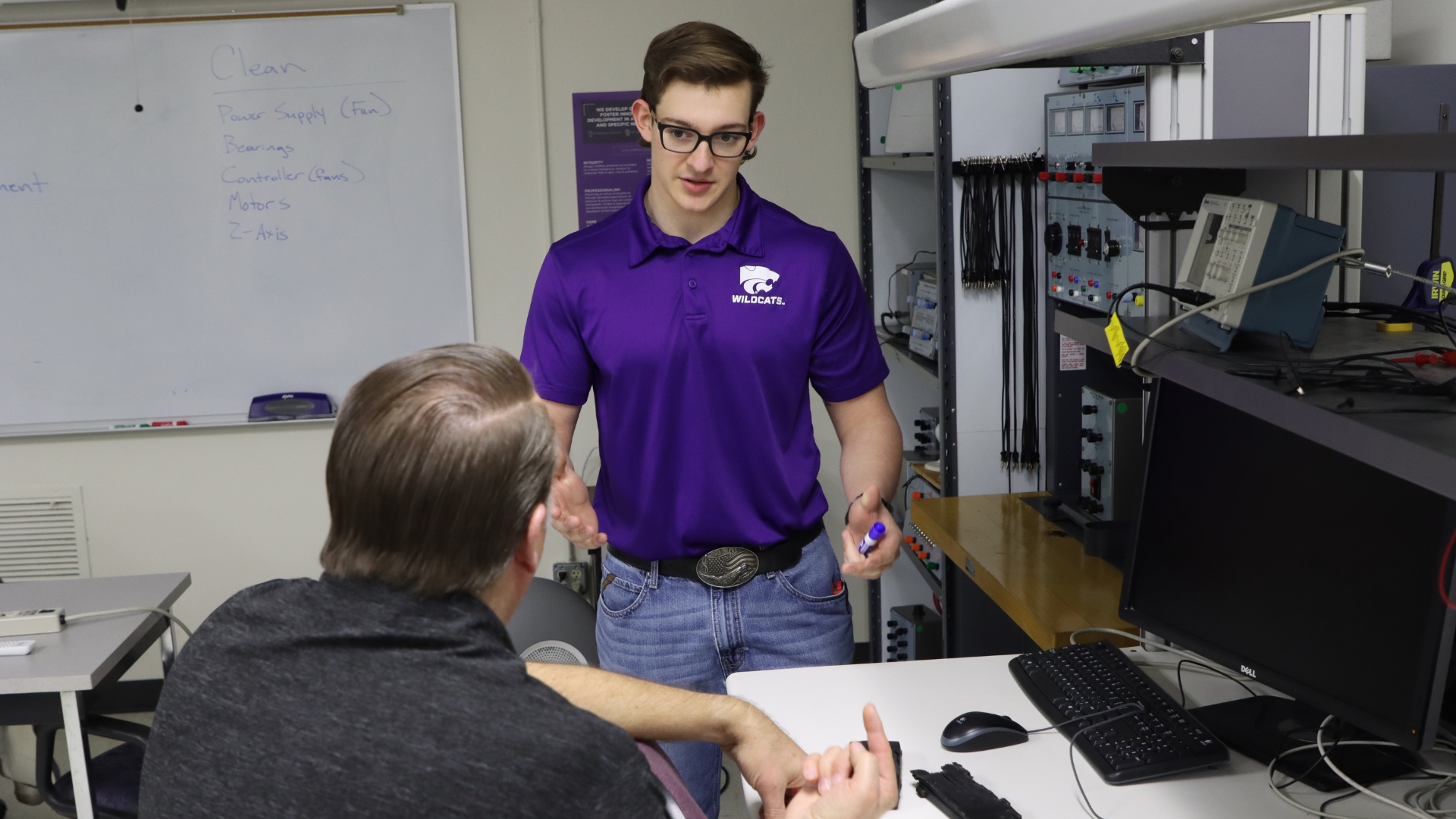 A student in a purple polo talks to one participant at a desk in the 3D printing cousrse