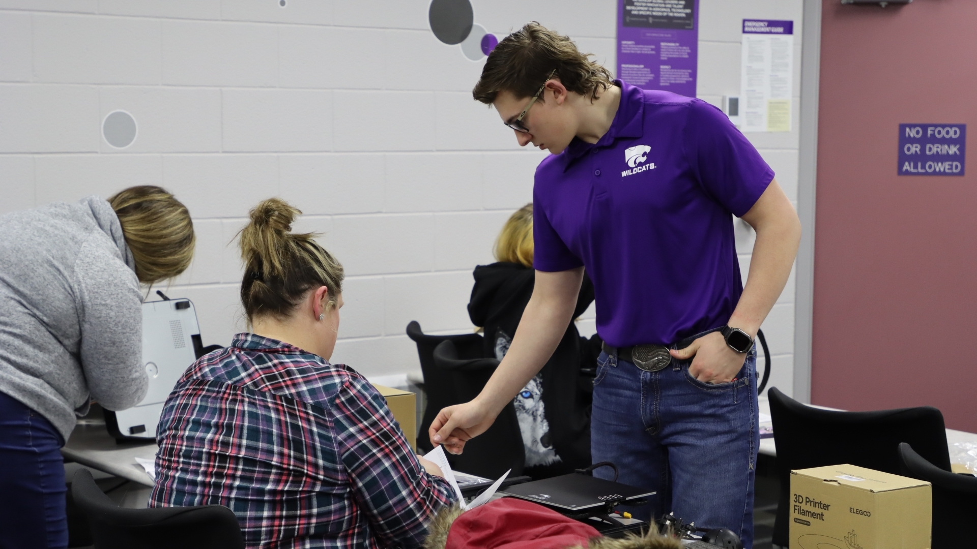 A student in a purple polo talks to participants seated at a table in a 3D printing course
