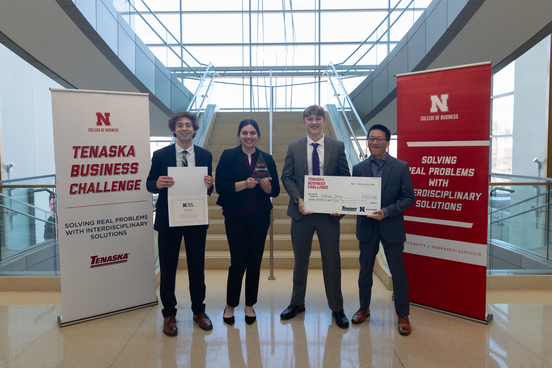 Four students in business attire stand between competition banners at the bottom of a stairway holding certificates
