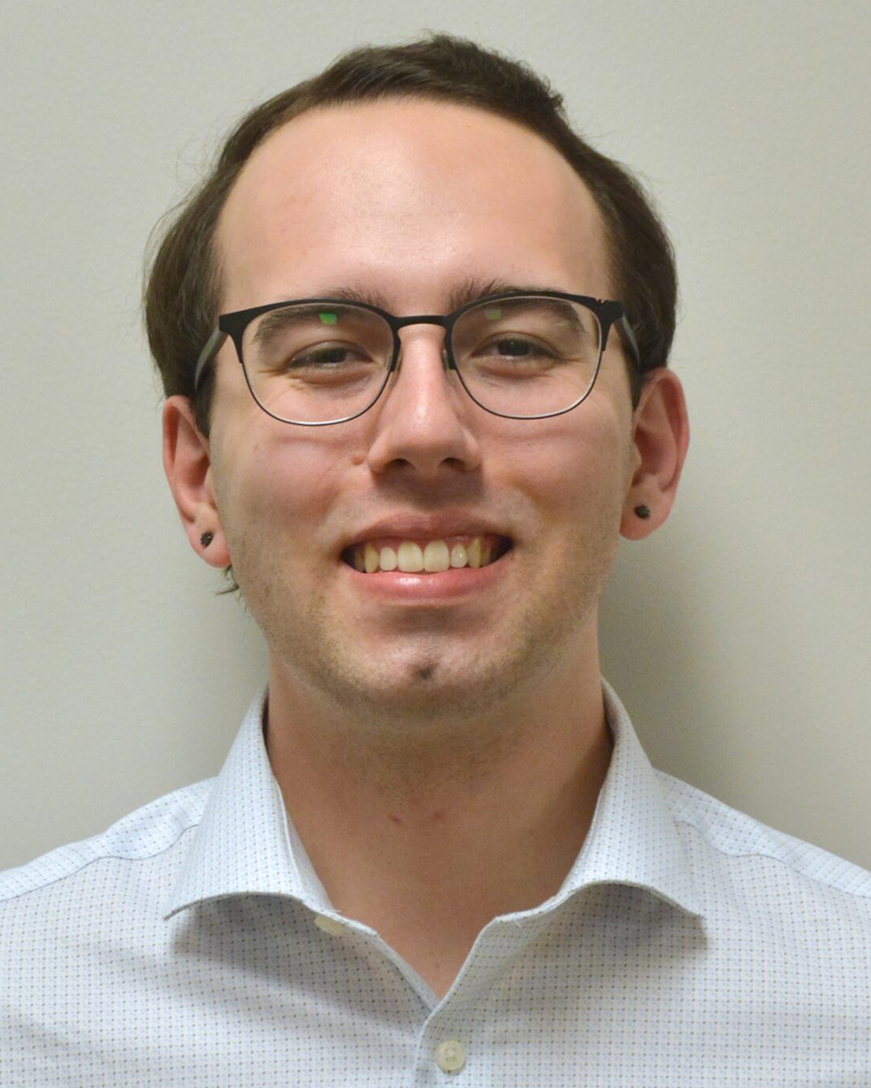 Man wearing a white button-up shirt and glasses smiles for a portrait against a white wall. 