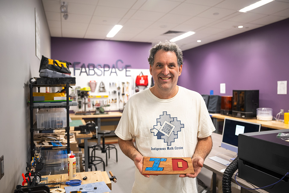 David Auckly stands in a workshop holding a carved wood sign that says ID.