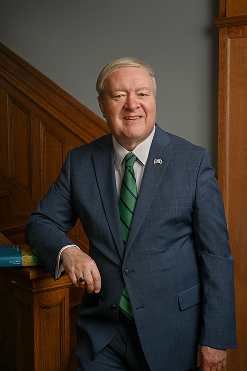 A gentleman in a dark blue-gray suit and a green tie leans against a wooden staircase bannister and smiles for a photo.