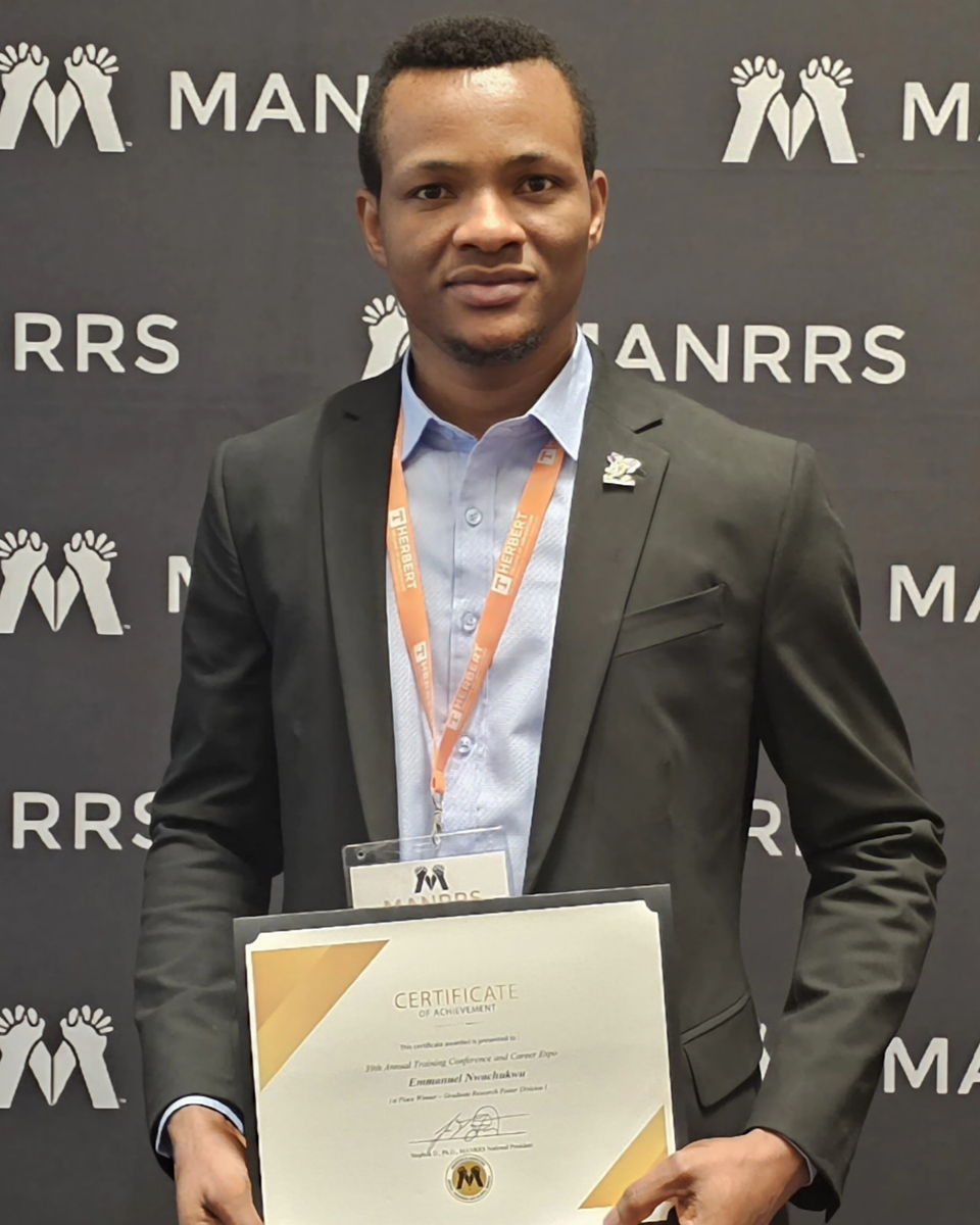 A master's student in a black blazer and light blue shirt holds a certificate in front of a blackdrop and poses for a portrait.