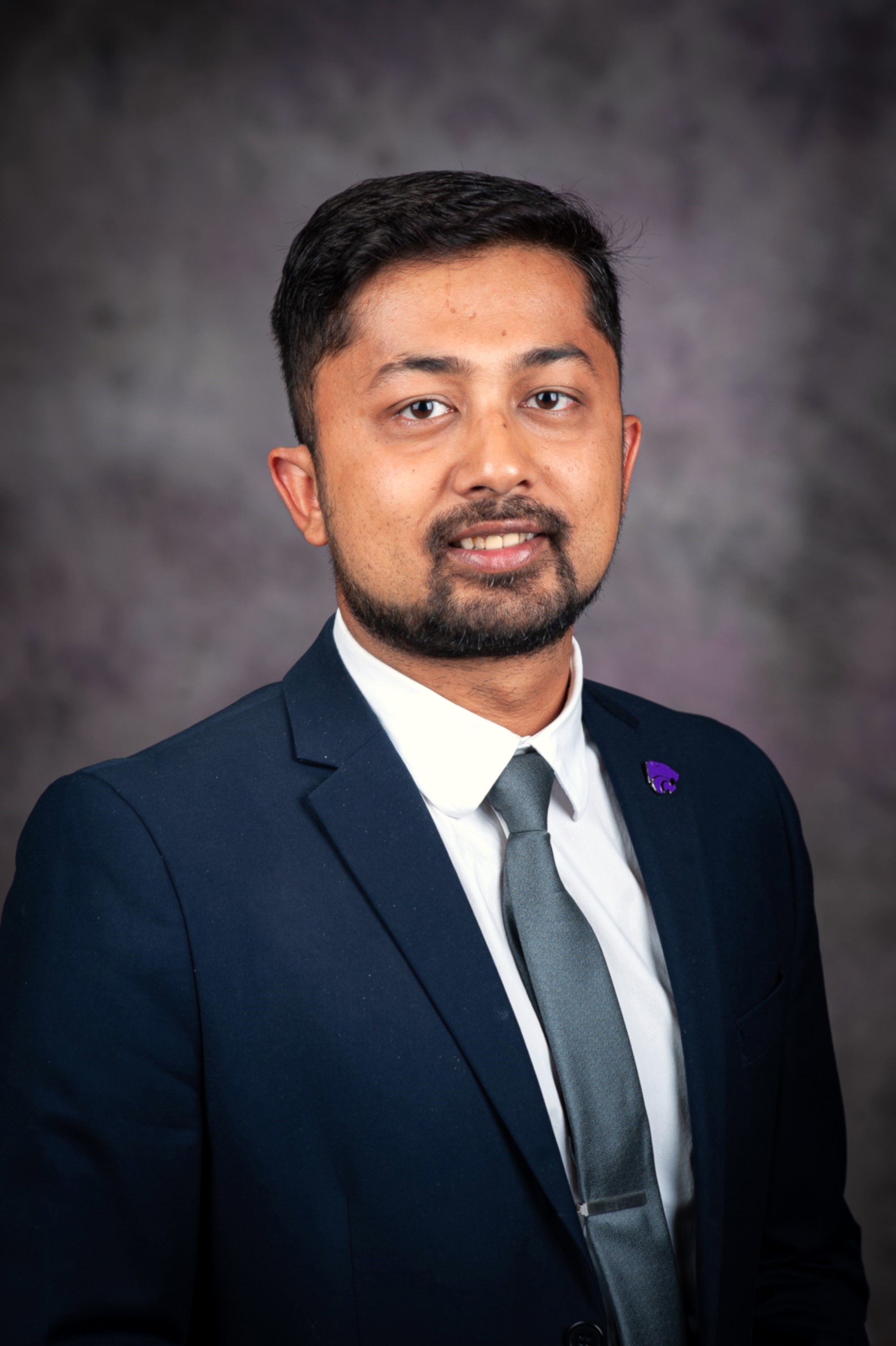 A man in a suit and tie with a powercat pin smiles for a portrait photo.