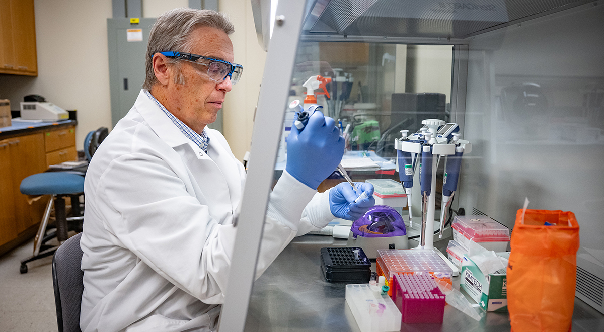 A man in a white lab coat and blue gloves with safety glasses works with tubes in a lab behind a protective hood