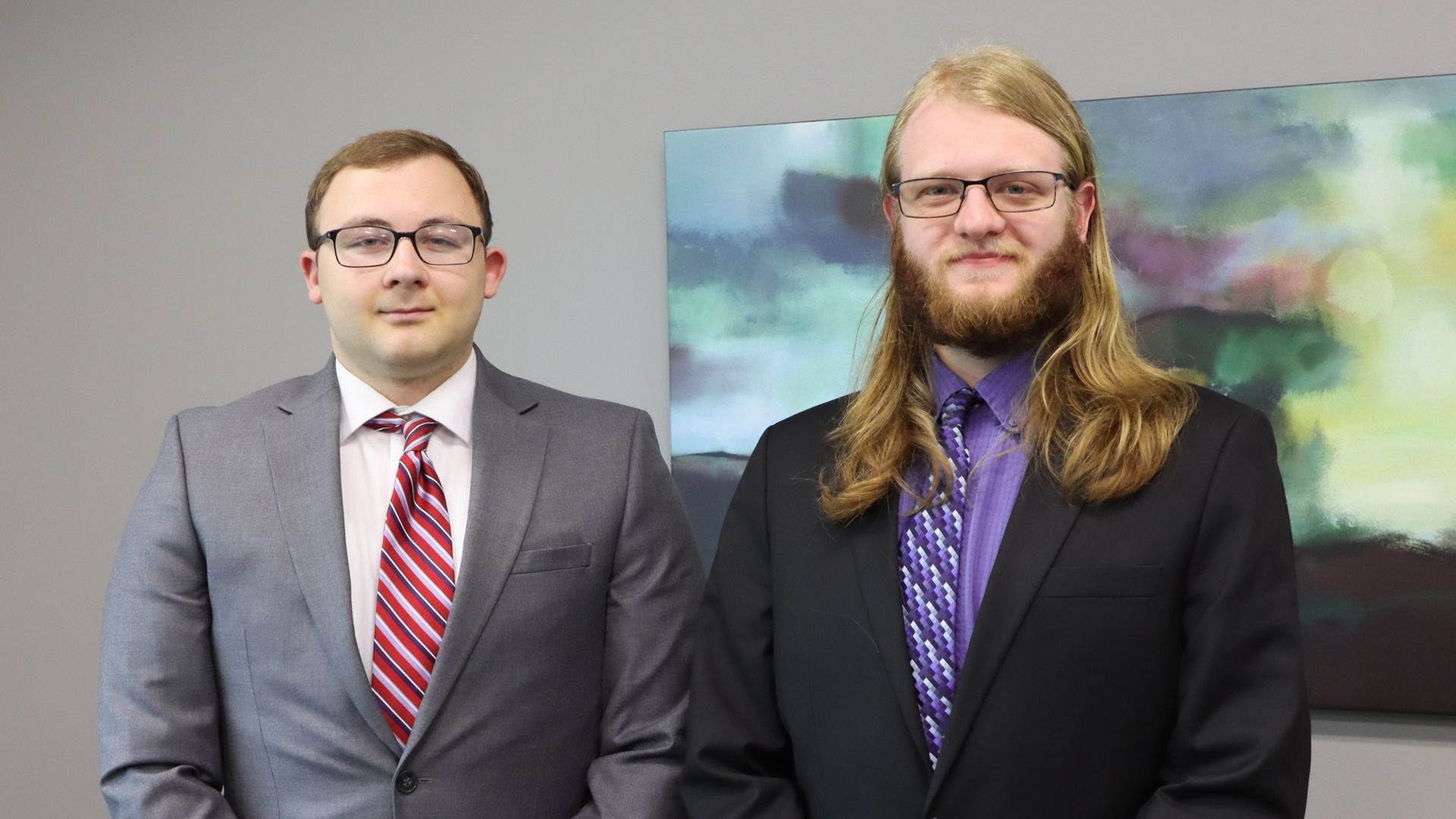 Two young men in suits and ties stand against a gray wall and smile for a photo.