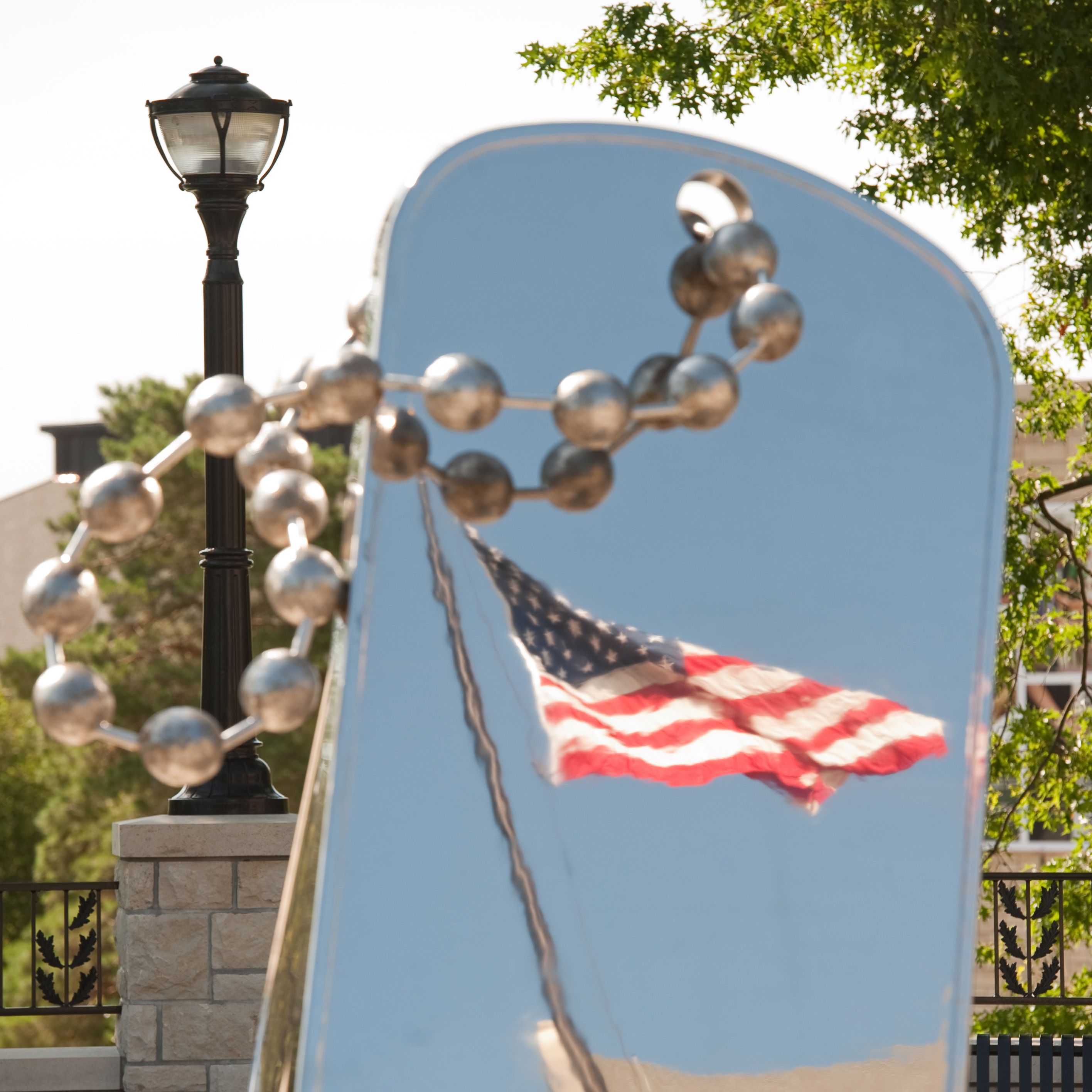 A waving American flag reflects against a clear blue sky on a large, silver and polished dog tag sculpture.