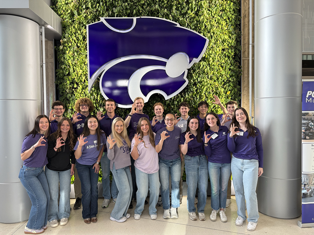 A group of students in purple shirts stand in front of a green wall covered in plants with a powercat on it.