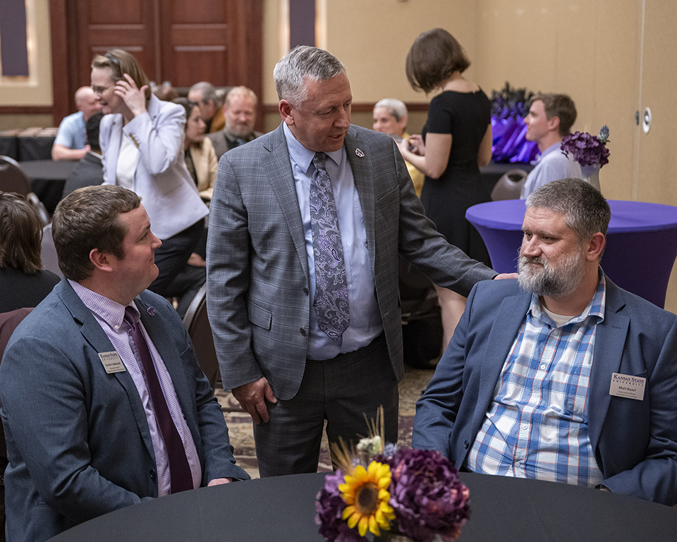 Two professors sit at a table while K-State President Richard Linton stands between them congratulates them.