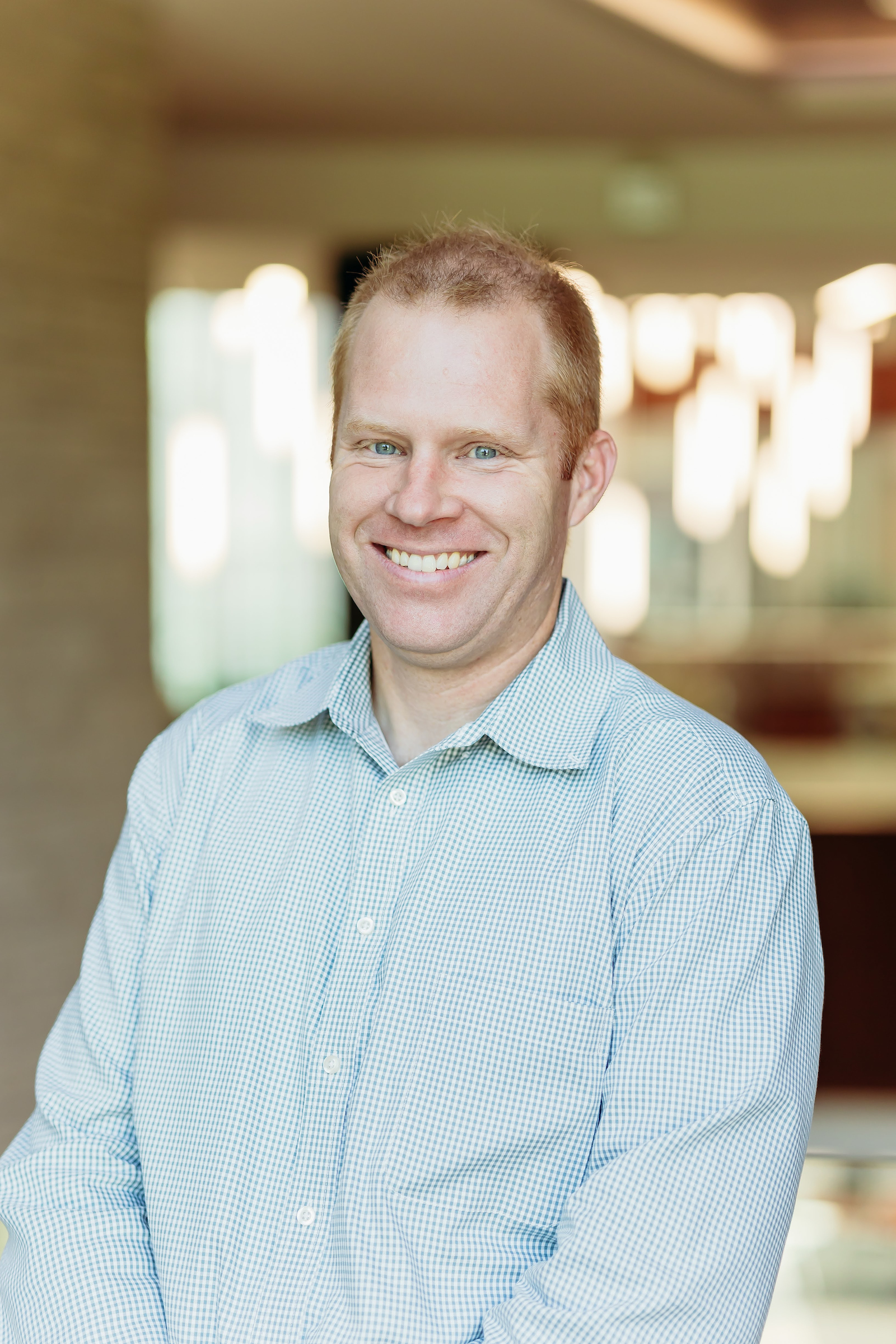 A white man with red hair in a blue and white checkered button-up shirt smiles for a portrait photo.