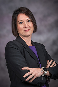 A woman with short brown hair in a suit with a purple shirt crosses her arms and smiles at the camera.