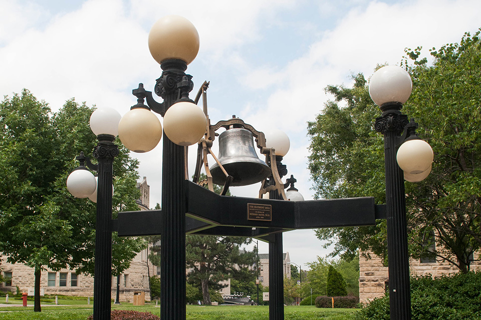 Against a blue sky and trees, a large metal beel is mounted on a metal structure with lamps. 