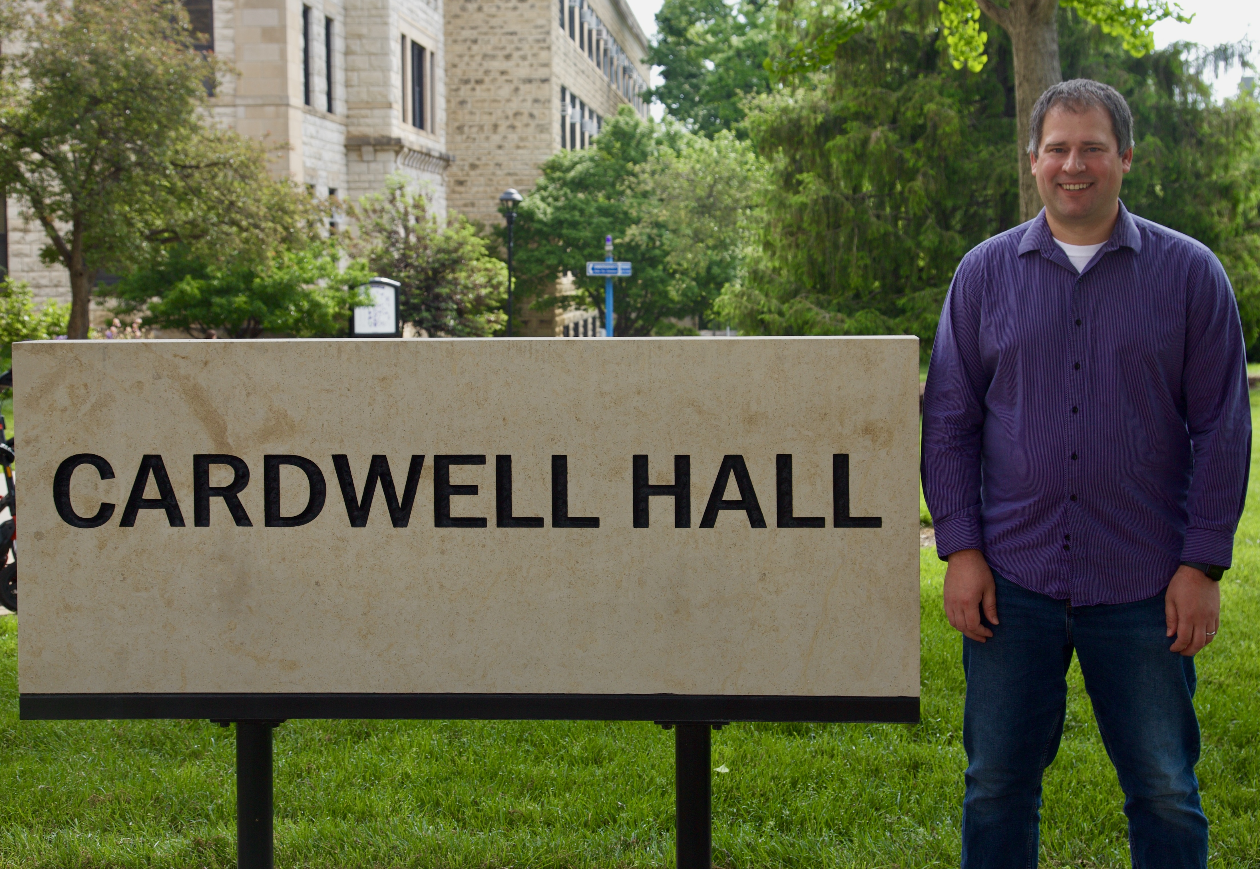 A man in a purple long-sleeved shirt smiling outside next to a Cardwell Hall limestone sign landscape photo.
