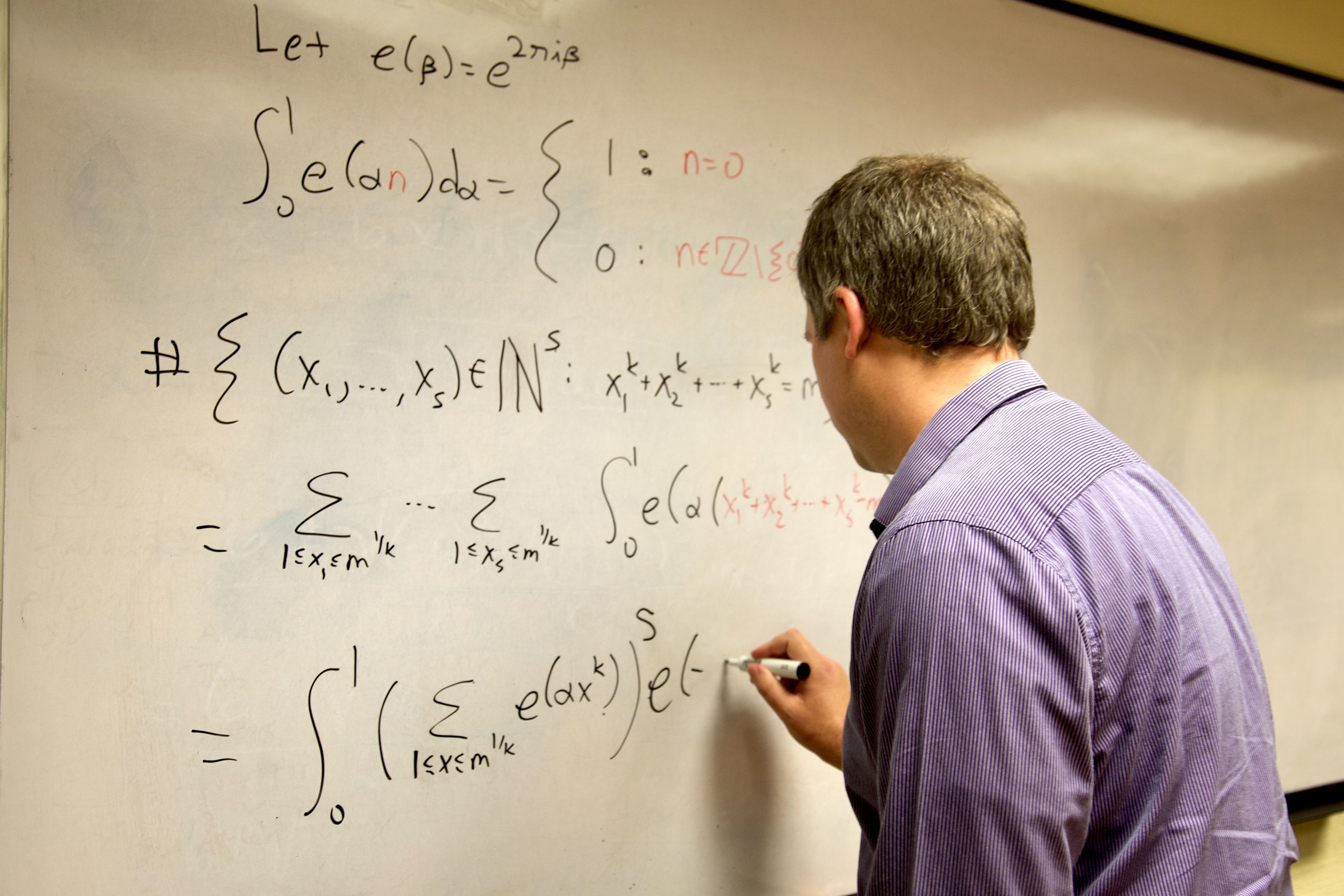 A man in a purple long-sleeved writing a math equation on a whiteboard.