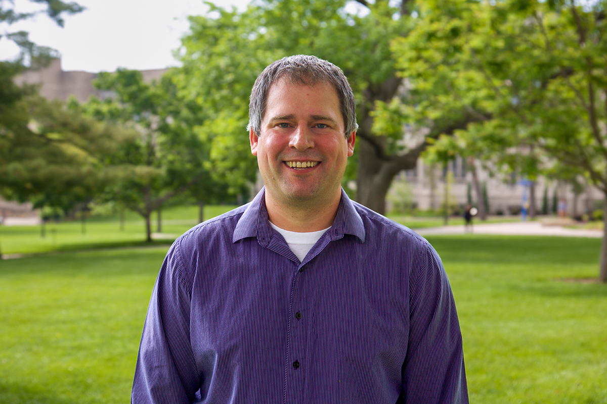 A man in a purple long-sleeved shirt smiling outside next to a Cardwell Hall limestone sign landscape photo.