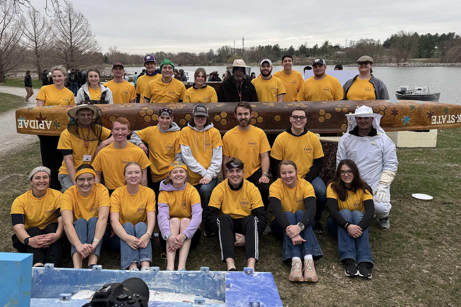 Several students and their professors pose for a group photo beside a lake and in front of the concrete canoe they designed and built.