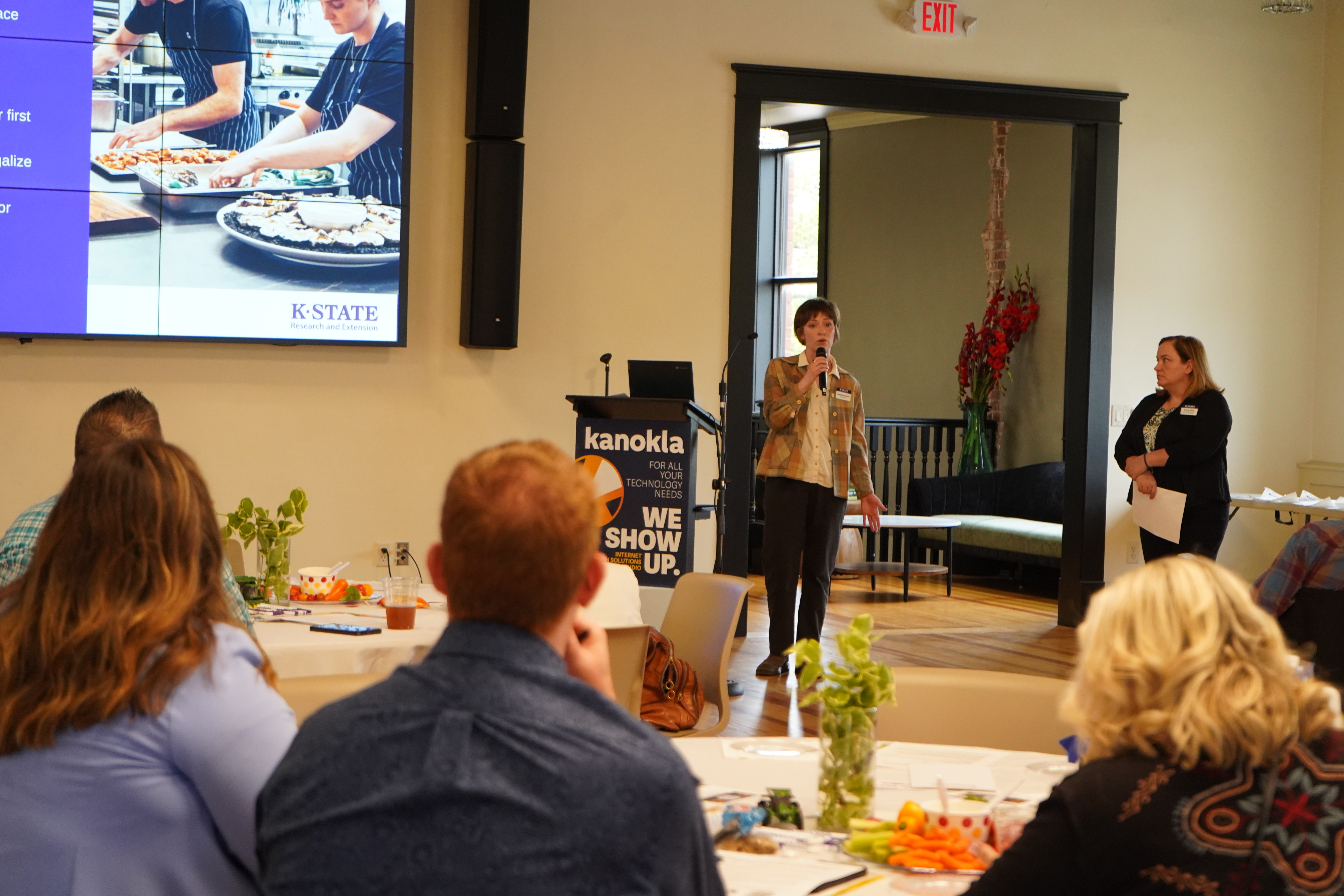 Two women stand at the front of a crowd giving a presentation that is shown on a large projector screen. 
