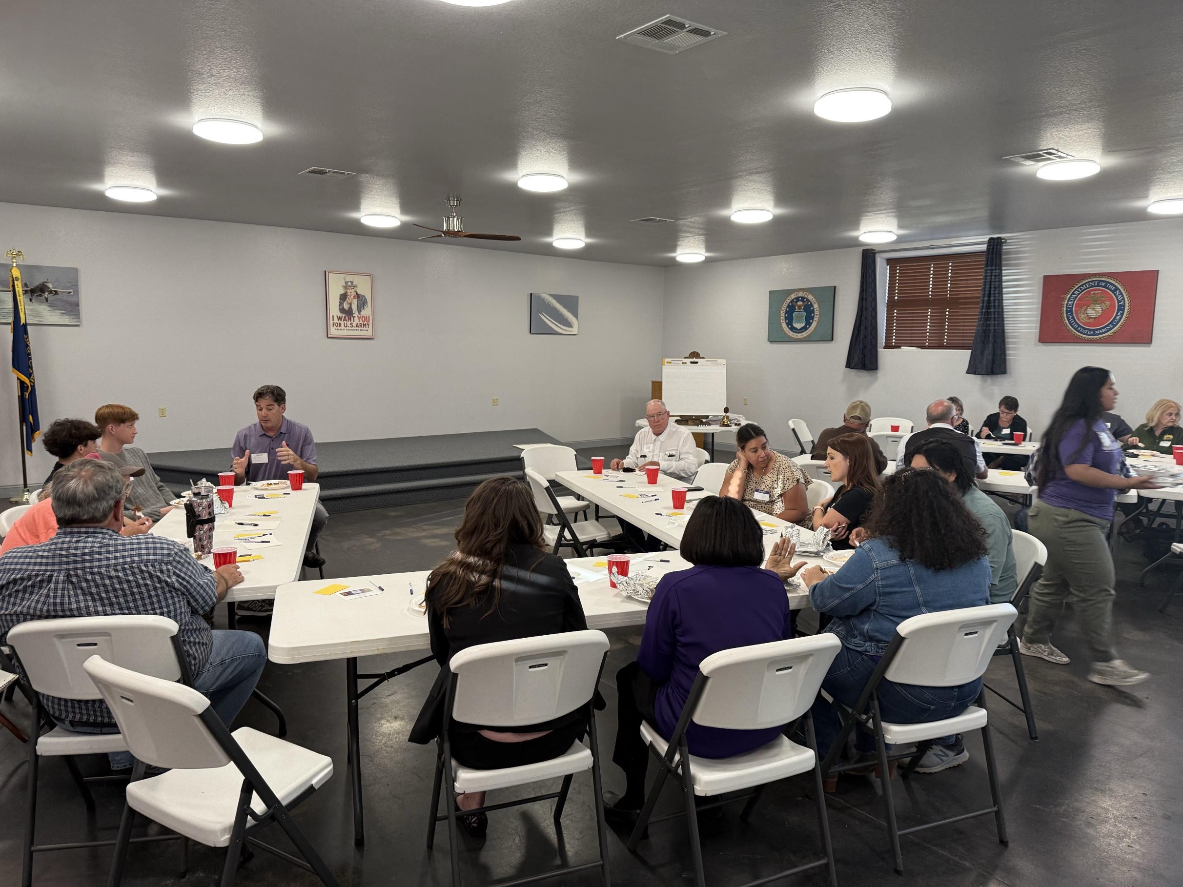 People are seated around white folding tables, engaging in a discussion.