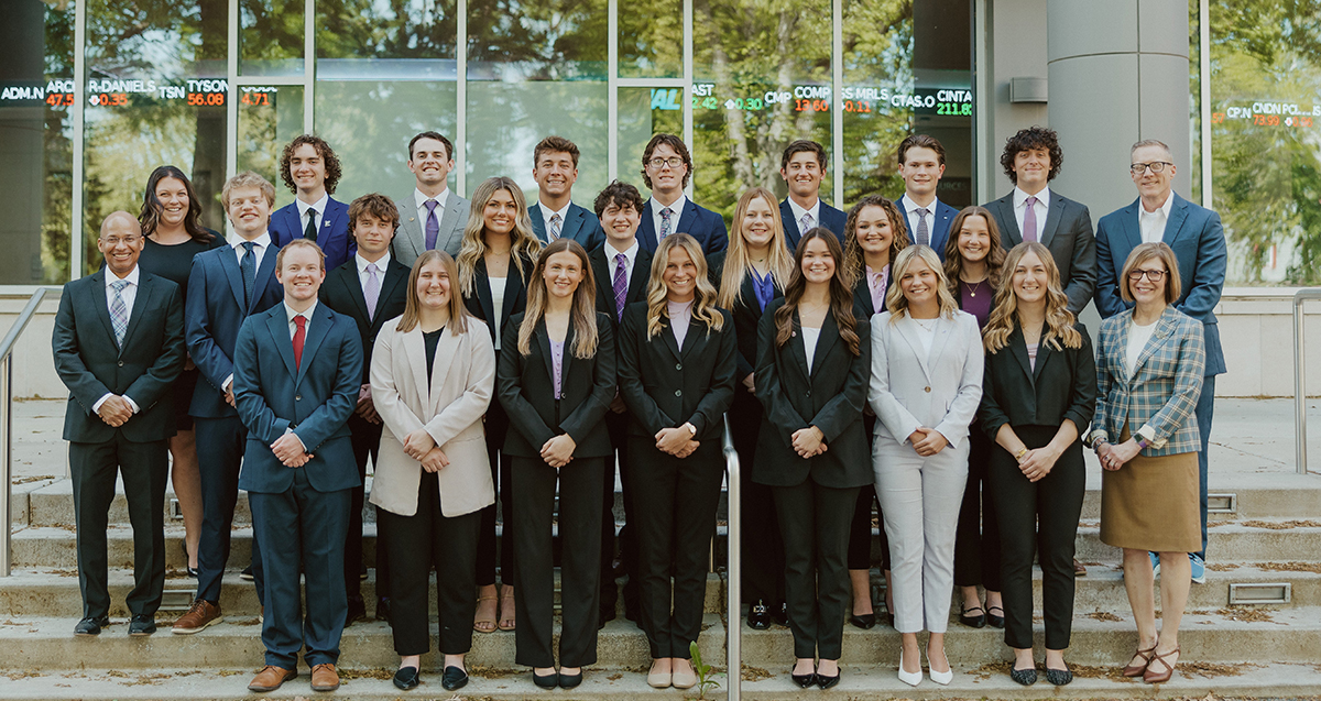 A group of students in professional attire posing for a group photo in front of glass windows.