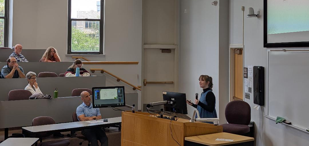 A student in a blue dress over a black long-sleeved turtleneck presents at the front of a room with a microphone to attendees.