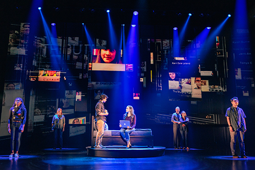 A stage play scene with actors under blue lighting in musical "Dear Evan Hansen." The background features projected images and text fragments. Central focus on two actors conversing with a laptop on a sofa, while others are positioned around the stage.