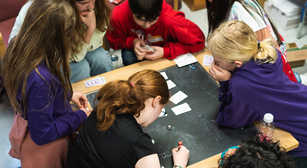 A group of young students gathered around a table with a black chalkboard on top.